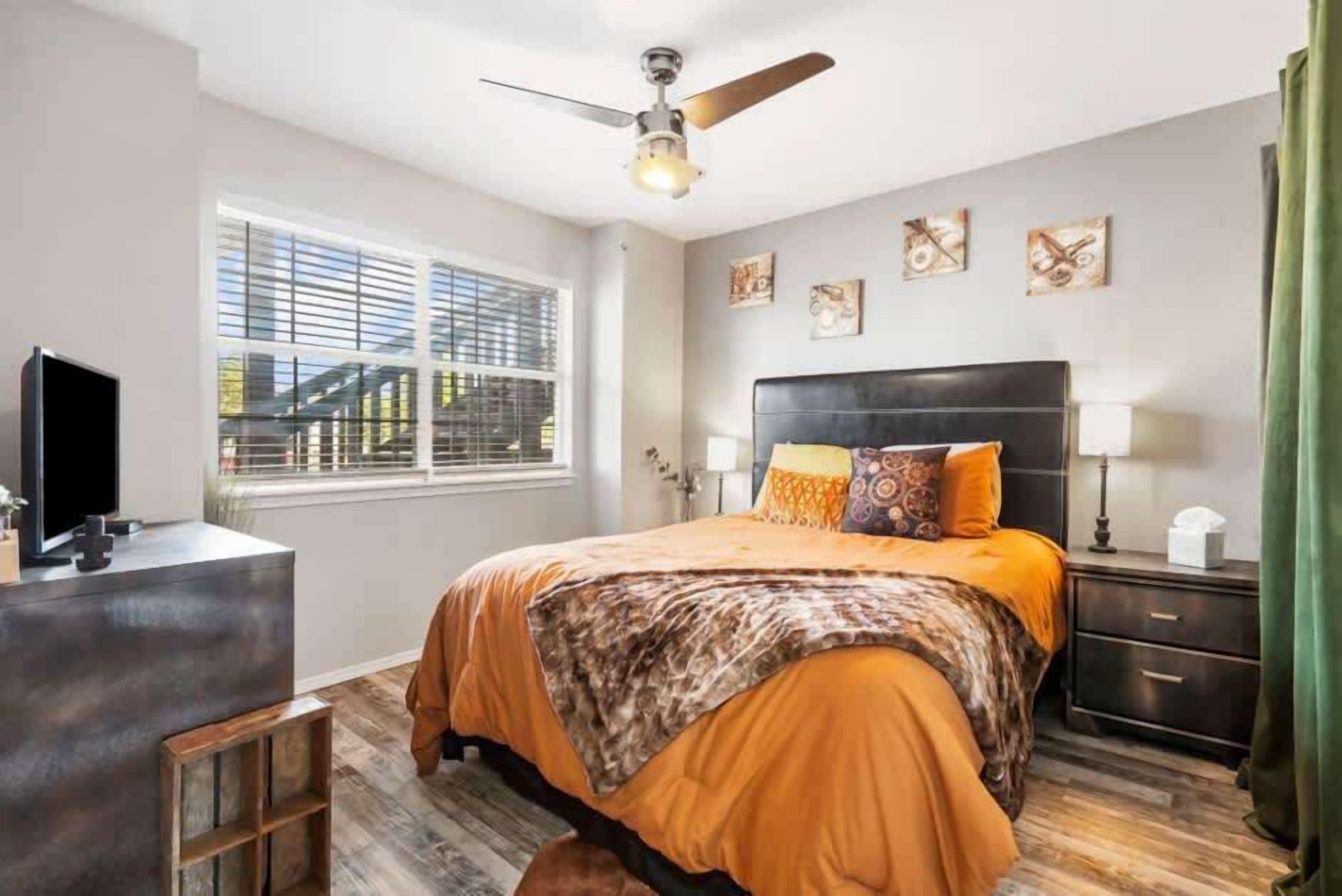 A bedroom with a bed, a desk, a ceiling fan, a window, and artwork on the wall.