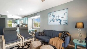 A modern living room with a sofa, two armchairs, a coffee table, and a rug. There is a kitchen in the background with cabinets and a window.
