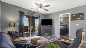 A living room with a sofa, ottomans, a chair, a fireplace, a ceiling fan, and a TV.