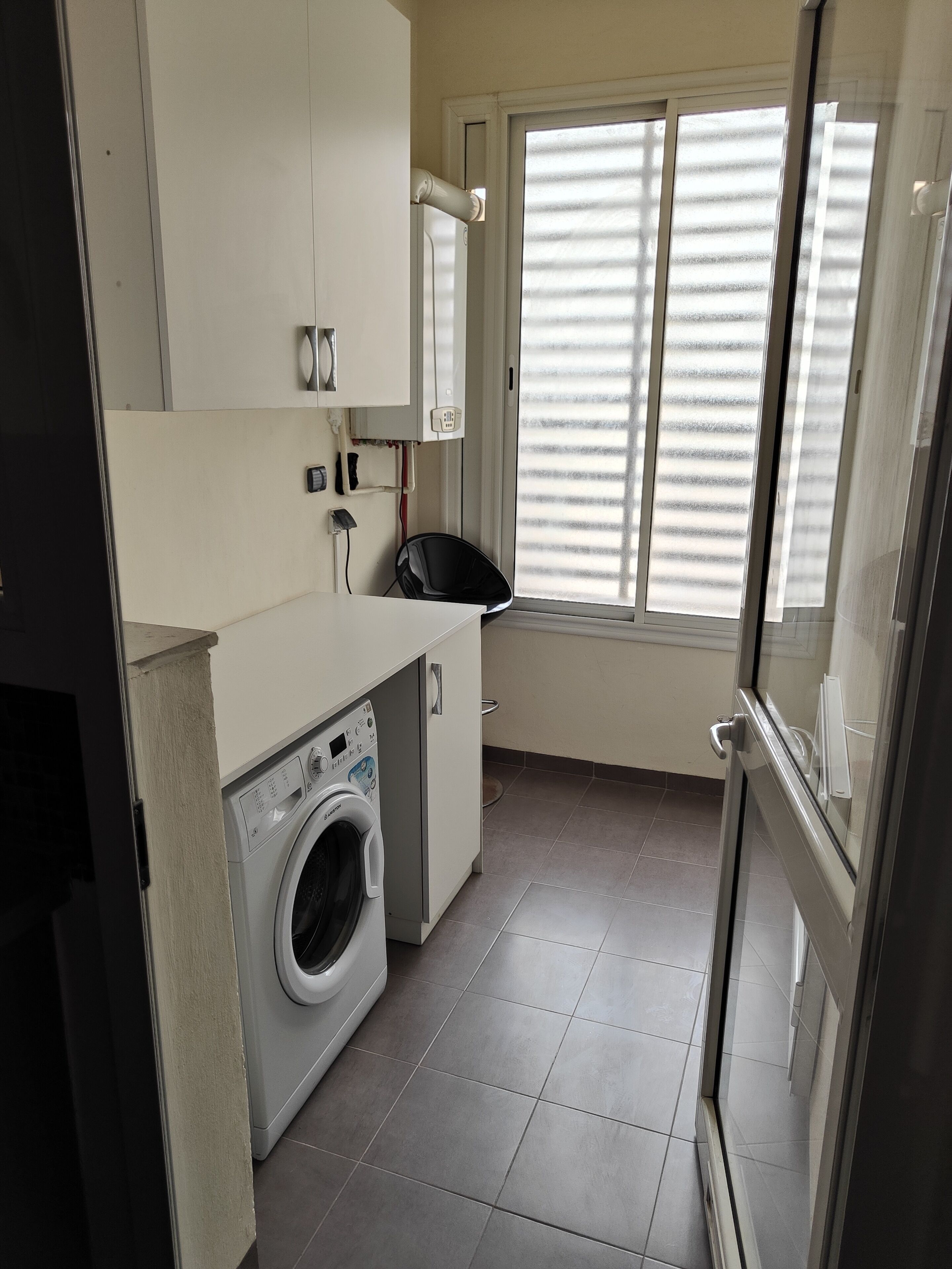 Une cuisine compacte dotée d’un lave-linge encastré, d’une fenêtre avec stores et d’un sol carrelé.