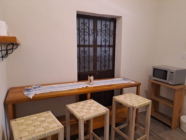 A small kitchen with a wooden table, four woven stools, a microwave on a shelf, and a decorative metal door.