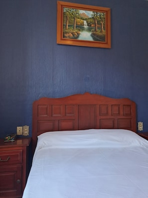A bedroom with a wooden headboard, a white bedspread, a nightstand with a lamp, and a painting of a landscape on the wall.