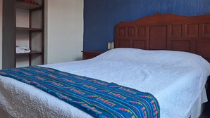 Une chambre à coucher avec une tête de lit en bois, une couvre-lit blanche et une couverture colorée. On y trouve une étagère en bois avec des vêtements pliés et une petite table avec une lampe.