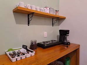 A kitchen counter with a coffee maker, a jar of sugar, a container of coffee, and a shelf with mugs and coffee containers.