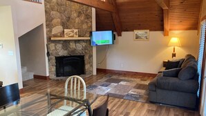 Ein gemütliches Wohnzimmer mit Kamin aus Stein, einem an der Wand befestigten Flachbildfernseher, einem bequemen grauen Sofa und einem Essbereich mit Glastisch und Stühlen.