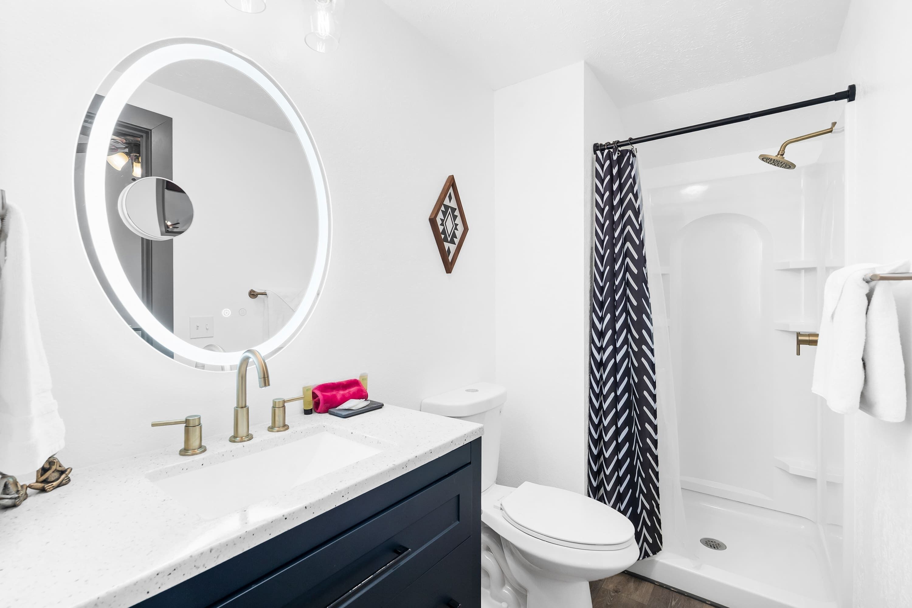 A modern bathroom with a round mirror, a white sink with gold fixtures, a toilet, and a shower with a black and white patterned curtain.