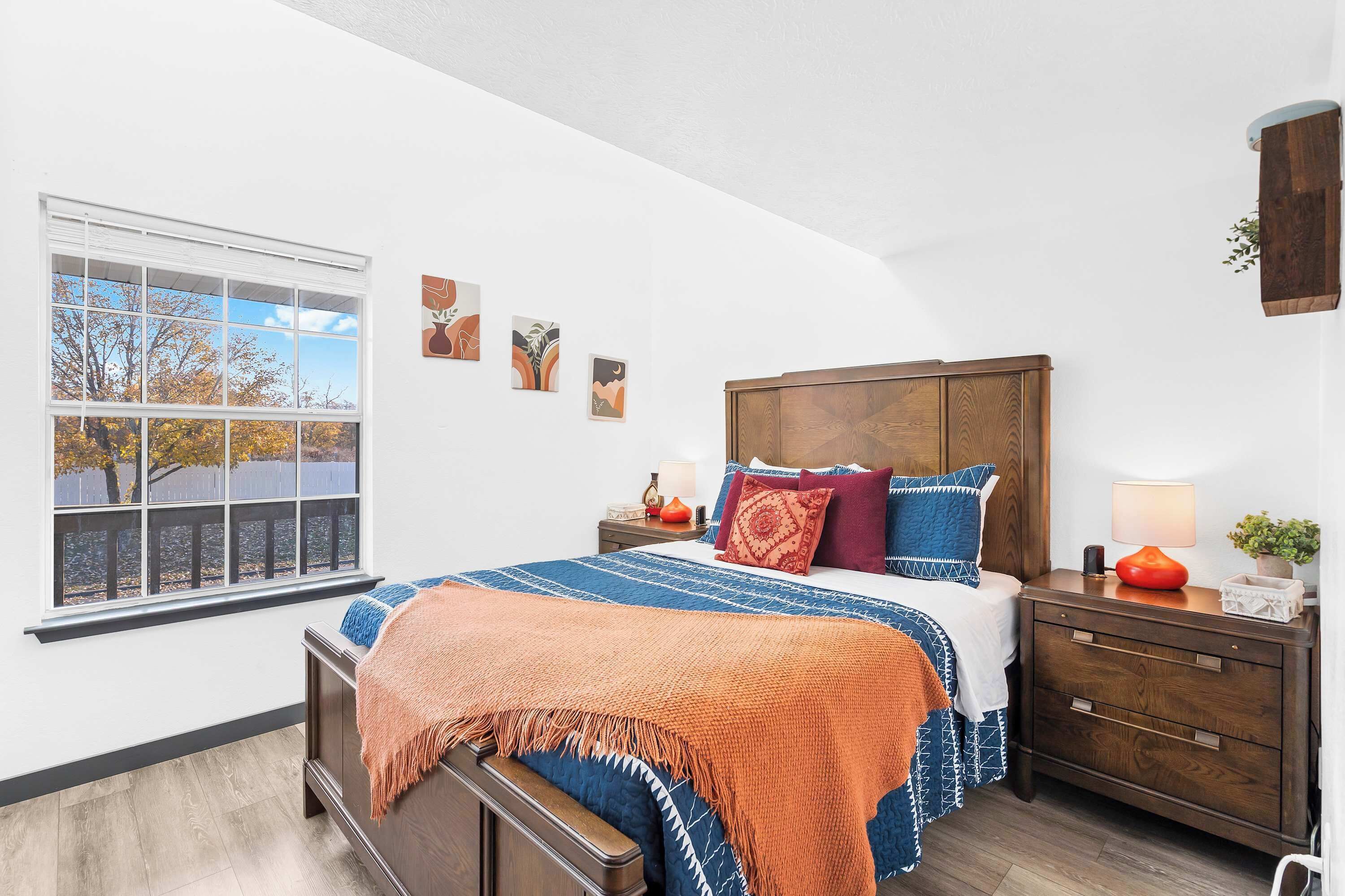 A bedroom with a large window, a wooden headboard, a bed with patterned bedding, a nightstand, and a wall-mounted shelf.