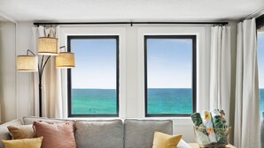 A modern living room with a grey sofa, yellow and brown cushions, and a small table with a basket of towels. Two large windows offer a view of the ocean. - SnowBirdsWelcome-GulfView-HeatedPool (Destin)