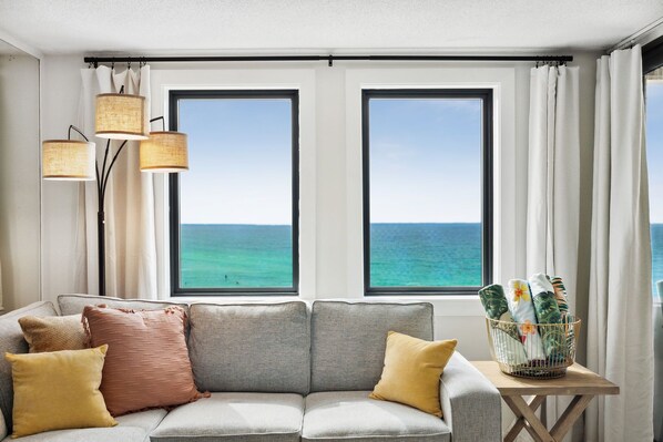 A modern living room with a grey sofa, yellow and brown cushions, and a small table with a basket of towels. Two large windows offer a view of the ocean. - SnowBirdsWelcome-GulfView-HeatedPool (Destin)