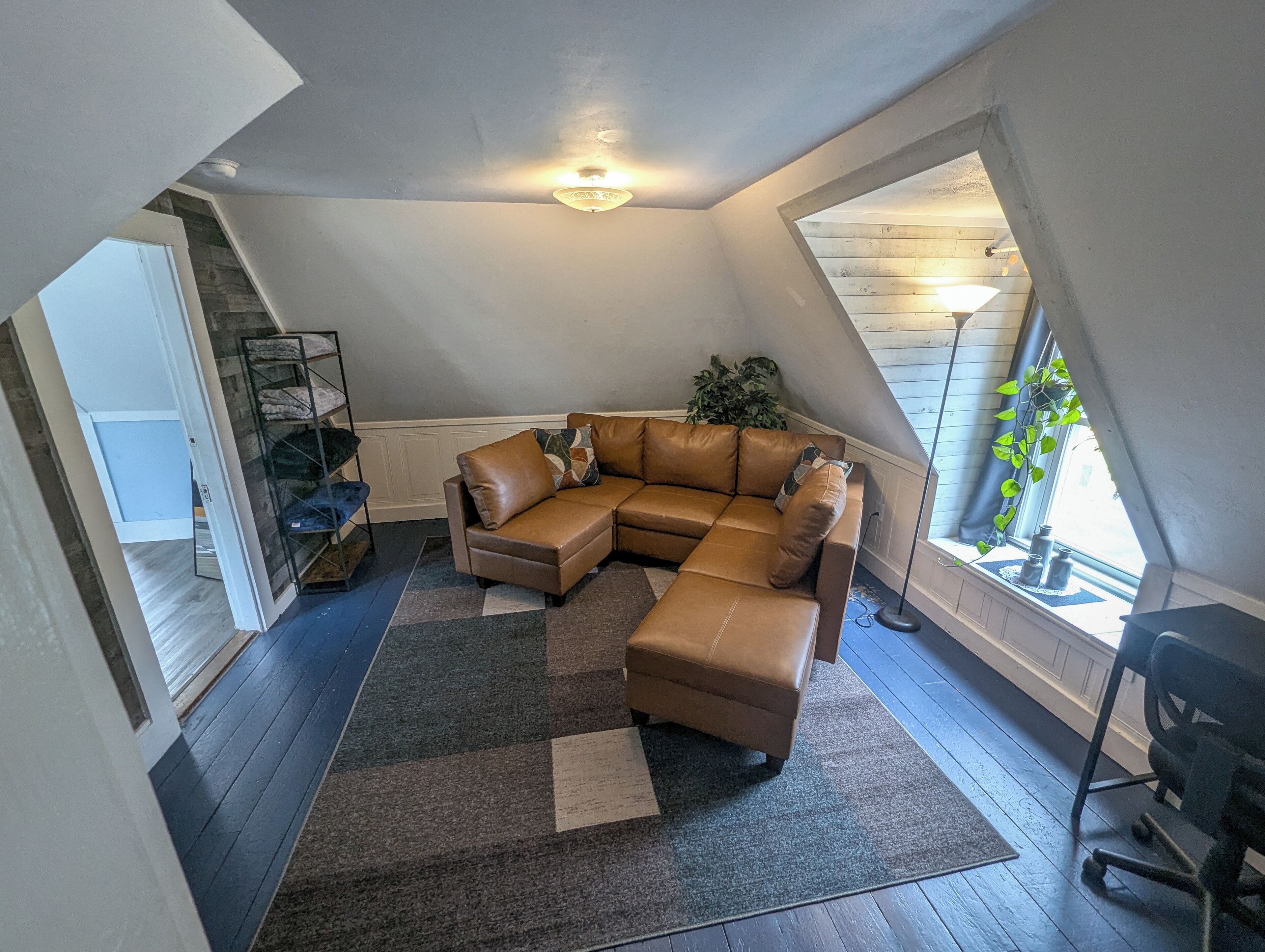 A loft-style living room with a brown leather sectional sofa, a small ottoman, and a dark patterned rug. There is a large window on the right side, a ceiling light, and a plant near the window.