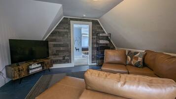 A cozy living room with a brown leather sofa, a flat-screen TV on a wooden stand, and a staircase leading to an upper level.
