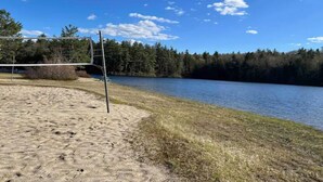 Une plage de sable où se trouvent un filet de volley-ball et une échelle, offrant une vue sur un lac paisible bordé d’arbres.