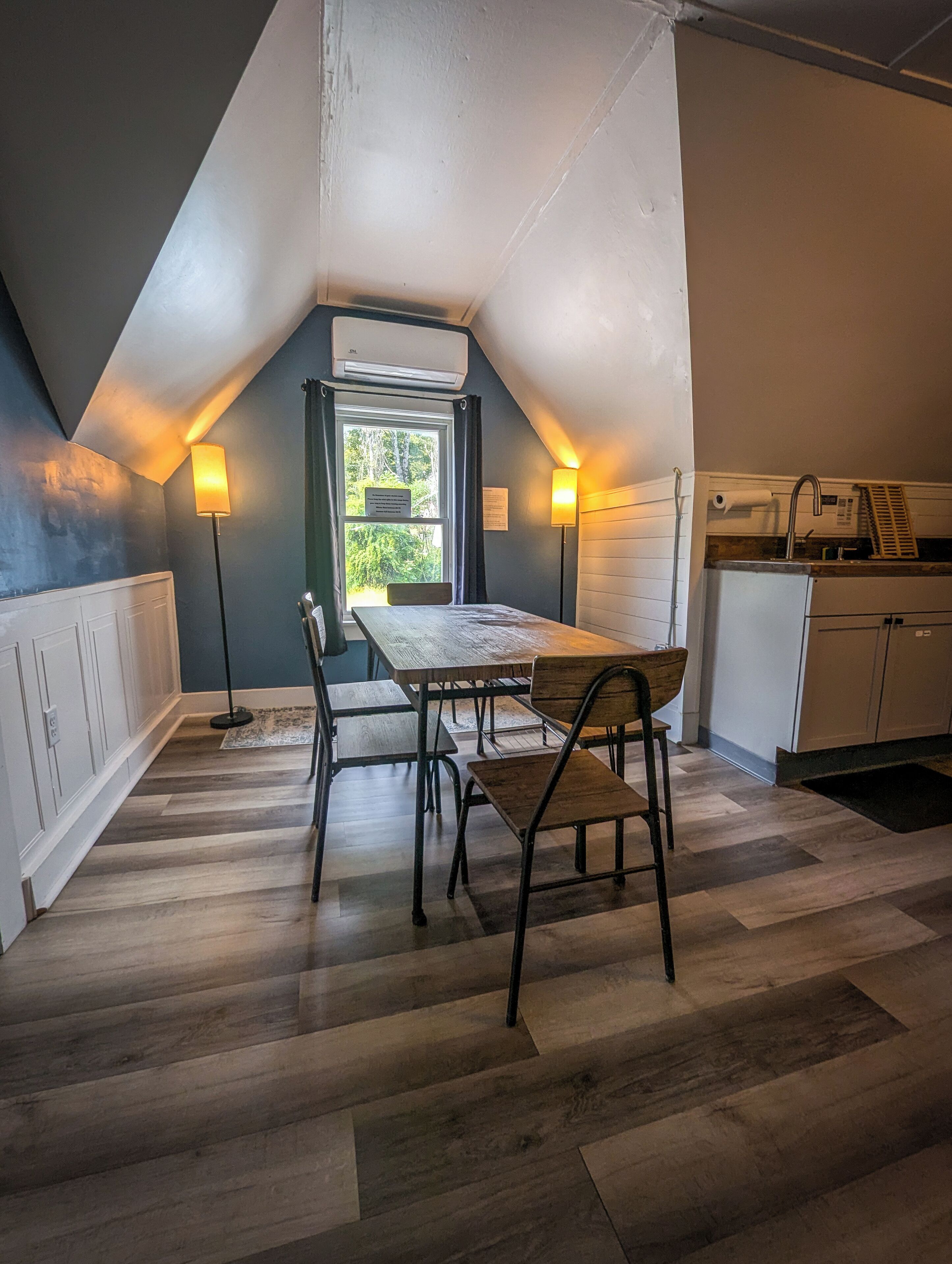 Een eethoek met een tafel en stoelen, een raam met uitzicht op groen, en een kitchenette met een koelkast en spoelbak.
