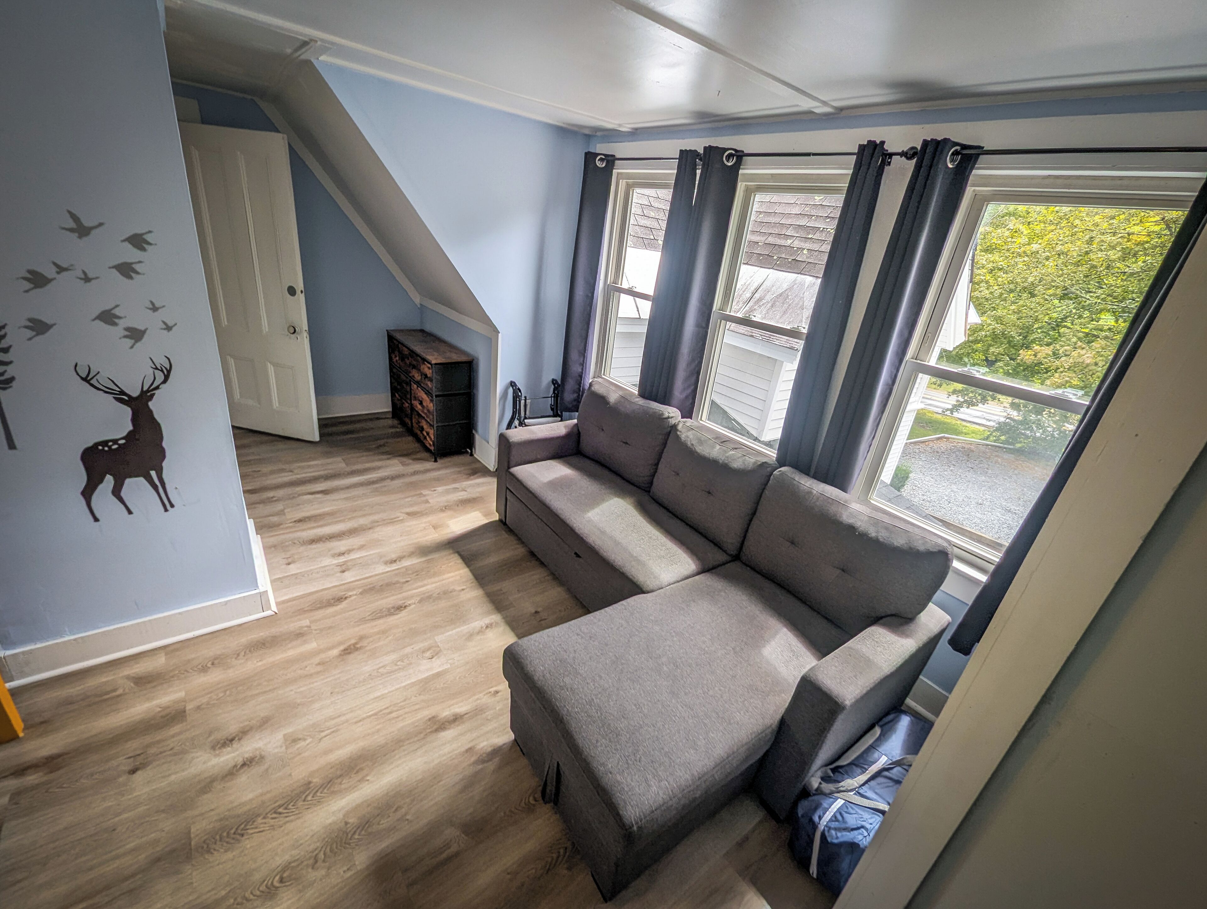 A room with a sectional sofa, a wooden floor, a wall decal of a deer, a staircase, and a window with curtains.