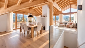 A spacious room with a wooden ceiling, a dining area with a table and chairs, a sofa, and a large window offering a mountain view.