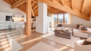 Un intérieur moderne comprenant une cuisine, un coin repas et un salon, agrémenté de poutres en bois, de rampes en verre et offrant une vue sur les montagnes.