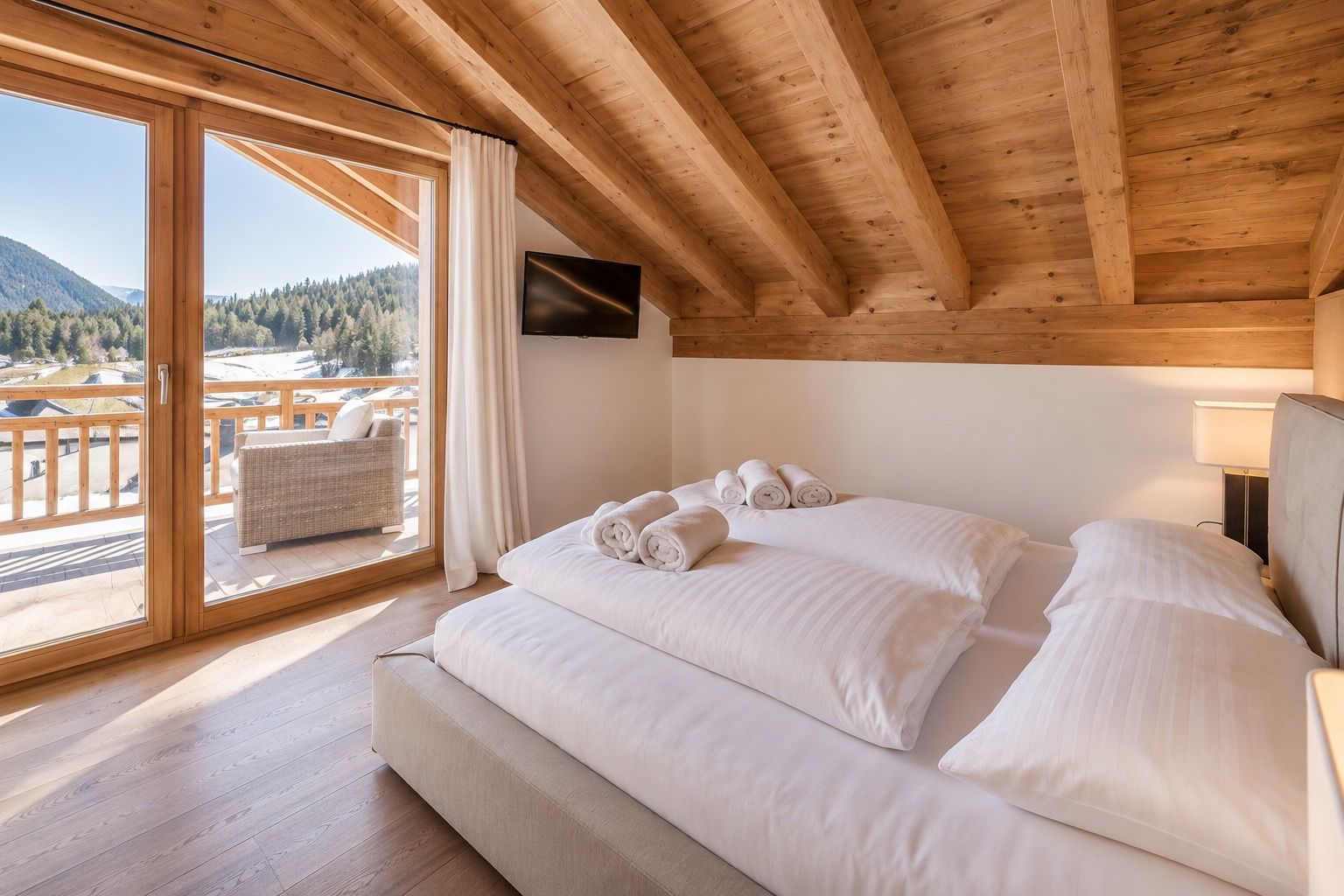 A bedroom with a large bed, a TV mounted on the wall, and a balcony with a view of a forested area.