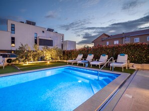 Ein Schwimmbecken mit Liegestühlen, einem gepflegten Rasen und modernen Gebäuden im Hintergrund.