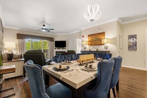 A dining area with a table set for four, blue upholstered chairs, a chandelier, and a television in the background.