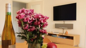 A dining table with a bottle, a vase of flowers, and a bowl of fruit. A flat-screen TV is mounted on the wall above a wooden cabinet. - Apartment "Meine Auszeit", 2 adults + 1 infant, 1 bathroom, terrace, WLAN (Koserow)