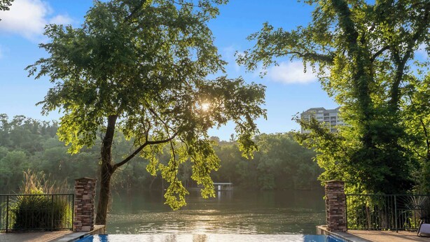 A swimming pool with a stone border, surrounded by trees and a view of a building across the water. - New Lakeview Penthouse - Amenities Galore! (Branson)