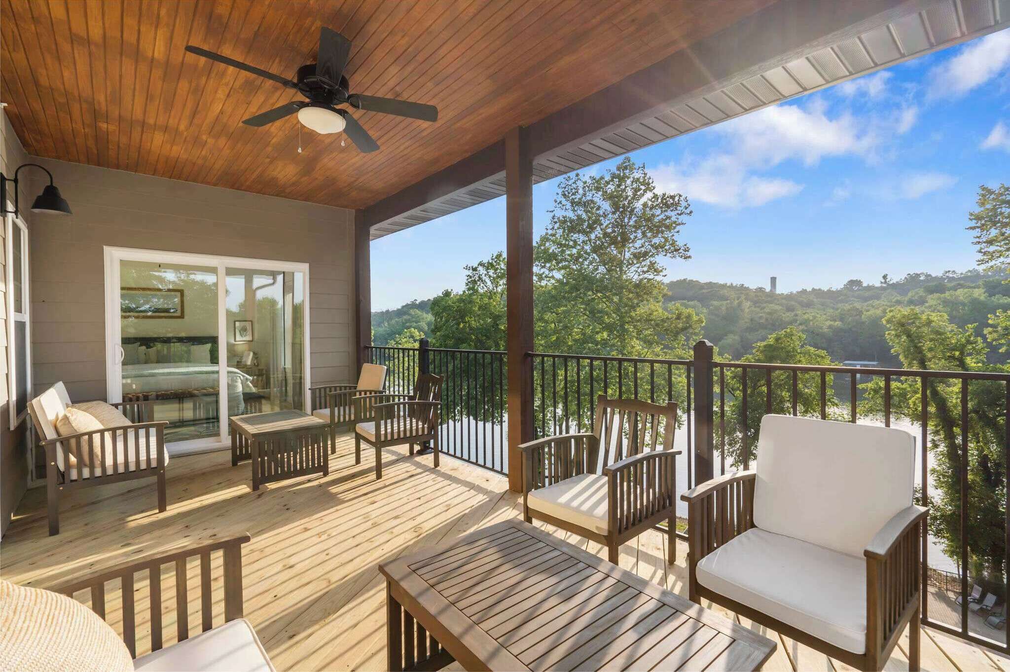 A covered deck with wooden furniture, a ceiling fan, and a view of a lake and forest.