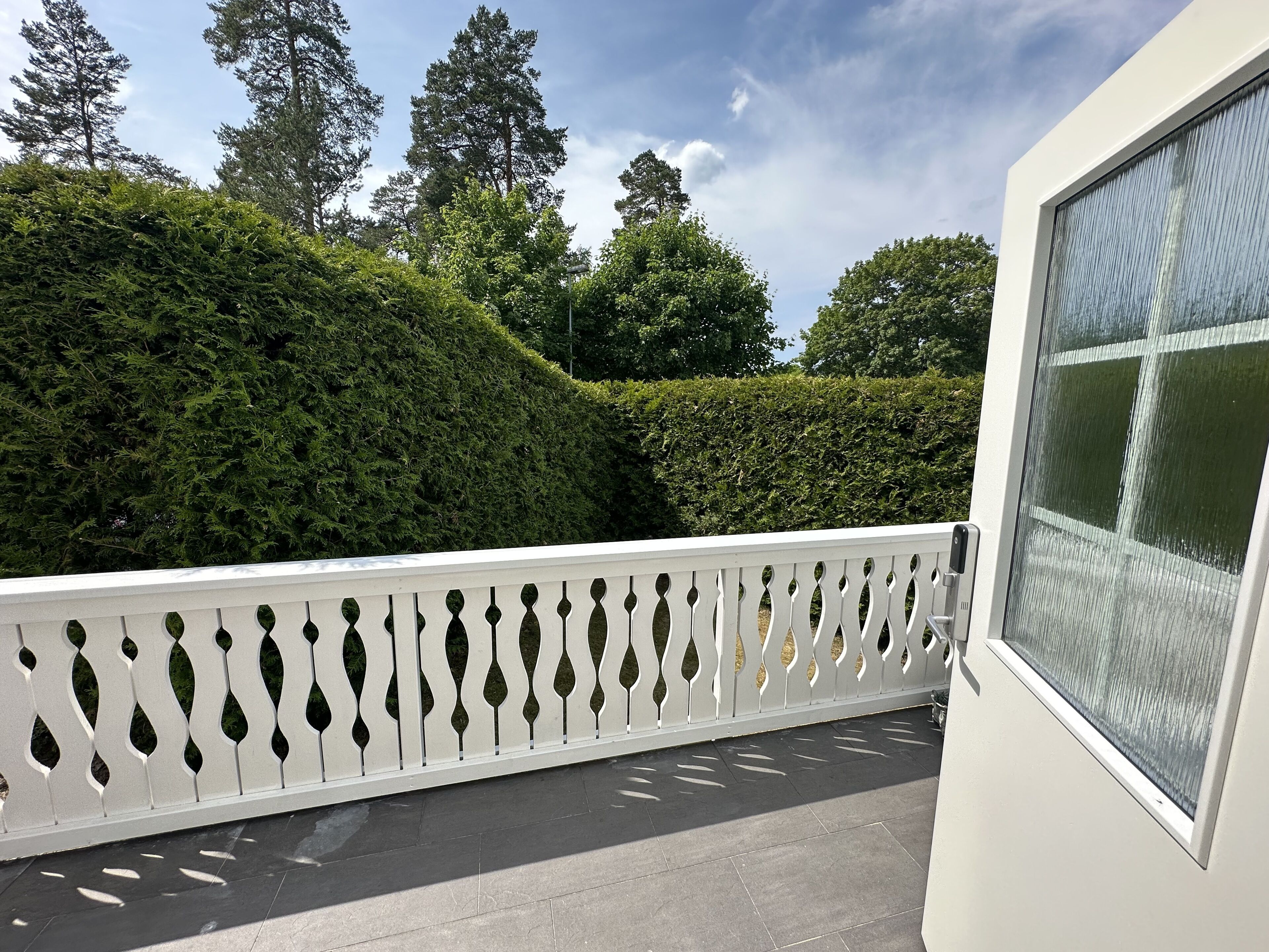 A balcony with a white decorative railing, a view of a well-maintained hedge, and tall trees in the background.