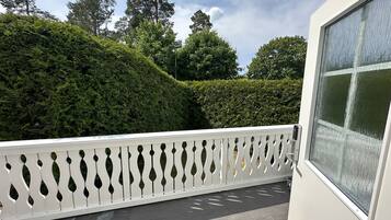 A balcony with a white decorative railing, a view of a well-maintained hedge, and tall trees in the background.