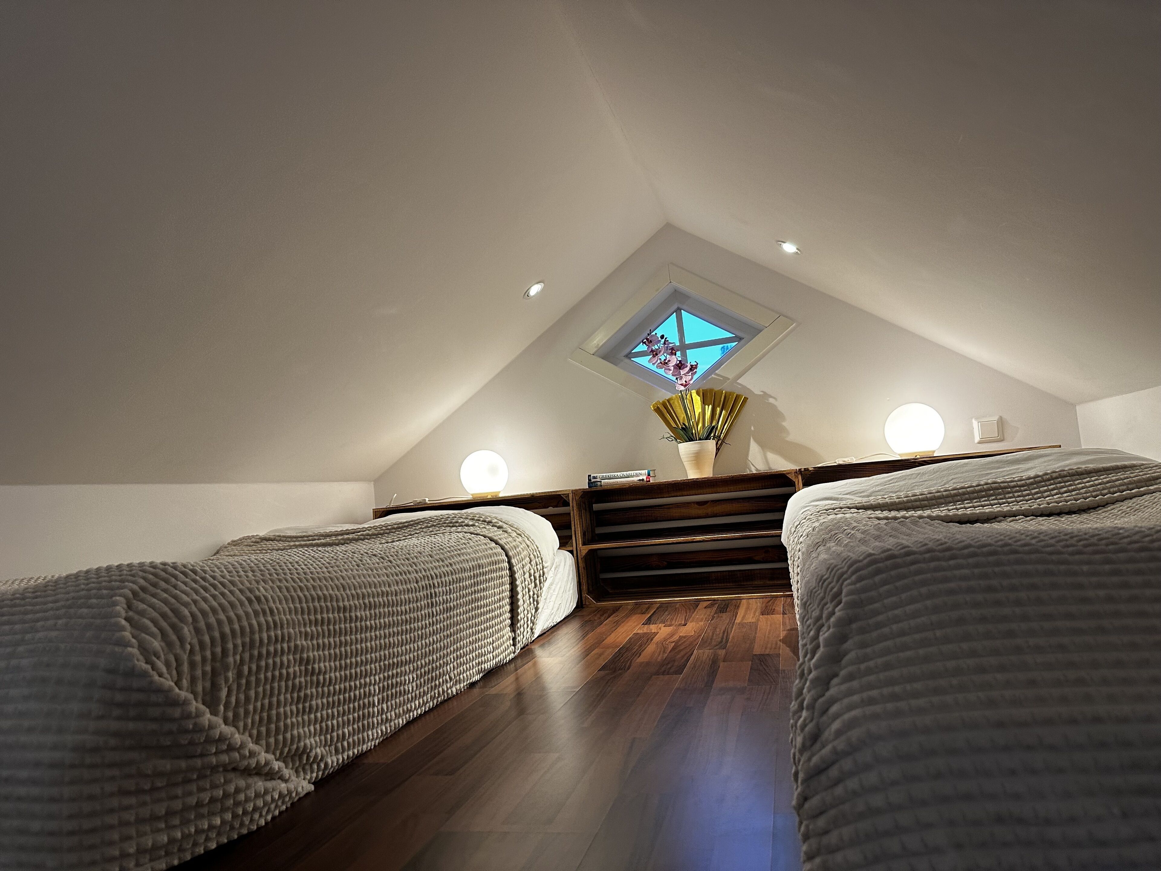 A cozy attic bedroom with two beds, a skylight, and a small table with a lamp.