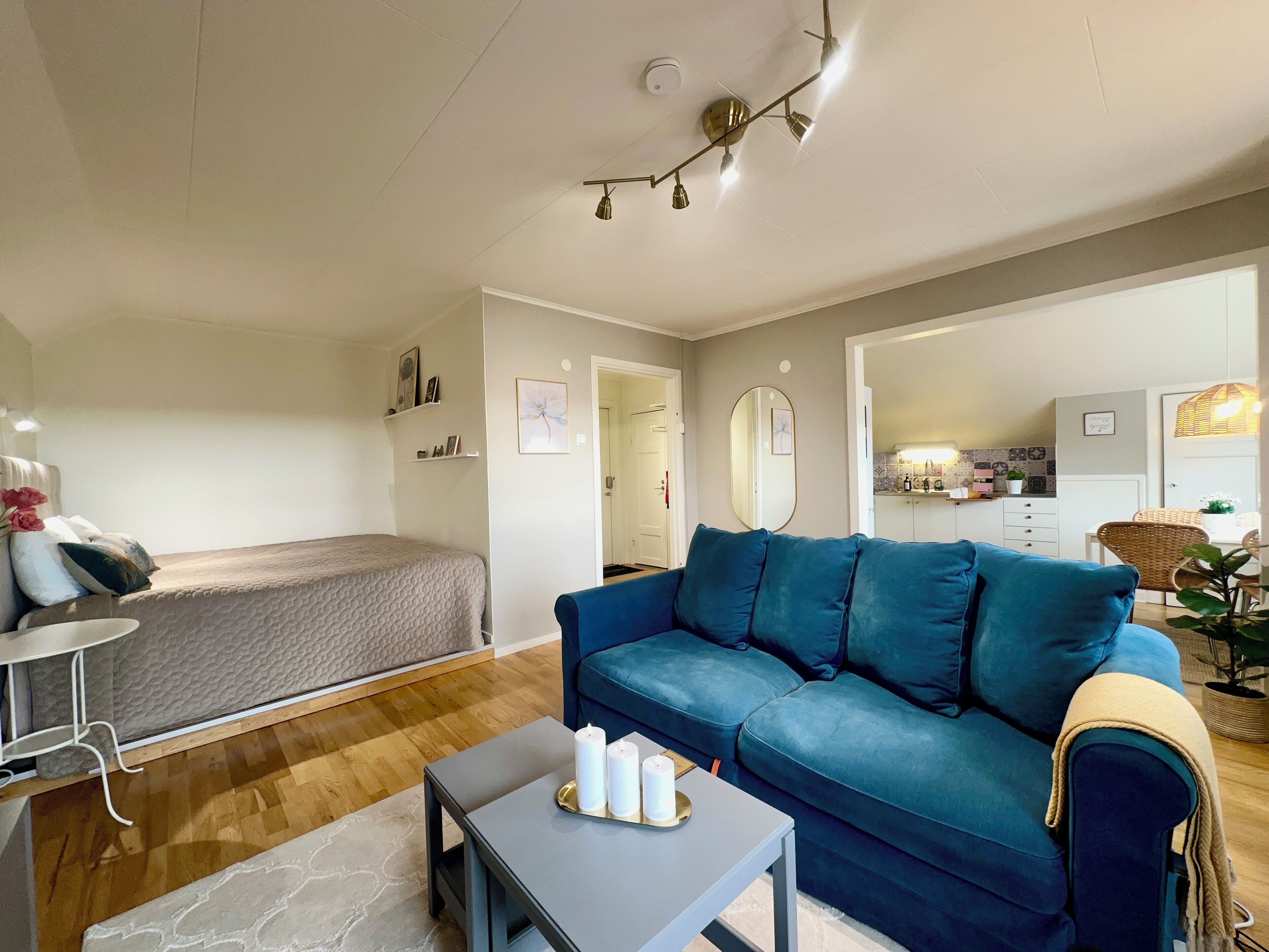 Uma sala de estar moderna com um sofá azul, uma mesinha com velas, uma cama e uma área de cozinha ao fundo.