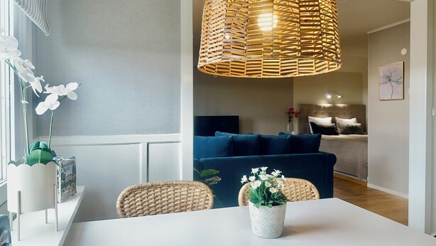 A dining area with a white table, wicker chairs, a wicker pendant light, and a potted plant with white flowers. - Prinshöjden Hotel - Apartments & Villas (Katrineholm)