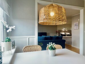 Un coin repas avec une table blanche, des chaises en osier, un luminaire suspendu en osier et une plante en pot ornée de fleurs blanches.