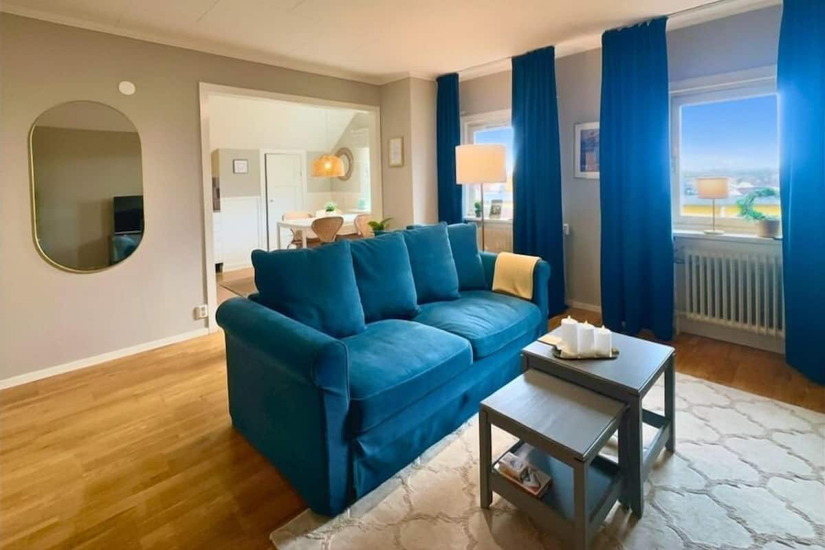 Ein Wohnzimmer mit einem blauen Sofa, einem kleinen Couchtisch, einem Spiegel und Blick ins Freie.