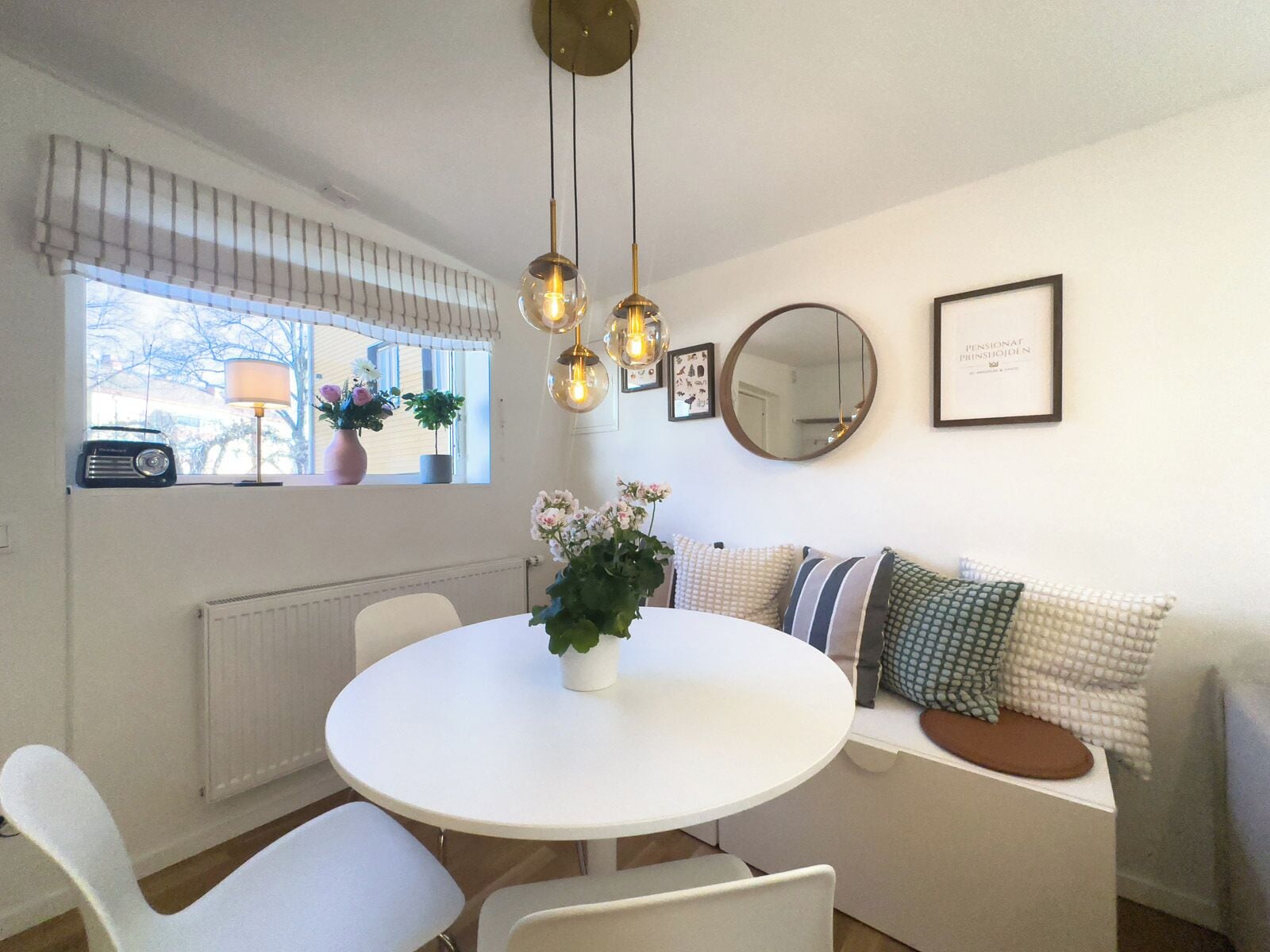 A dining area with a round table, white chairs, a radiator, a window with a radio, and a sofa with cushions.