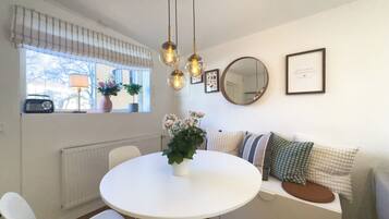 A dining area with a round table, white chairs, a radiator, a window with a radio, and a sofa with cushions.