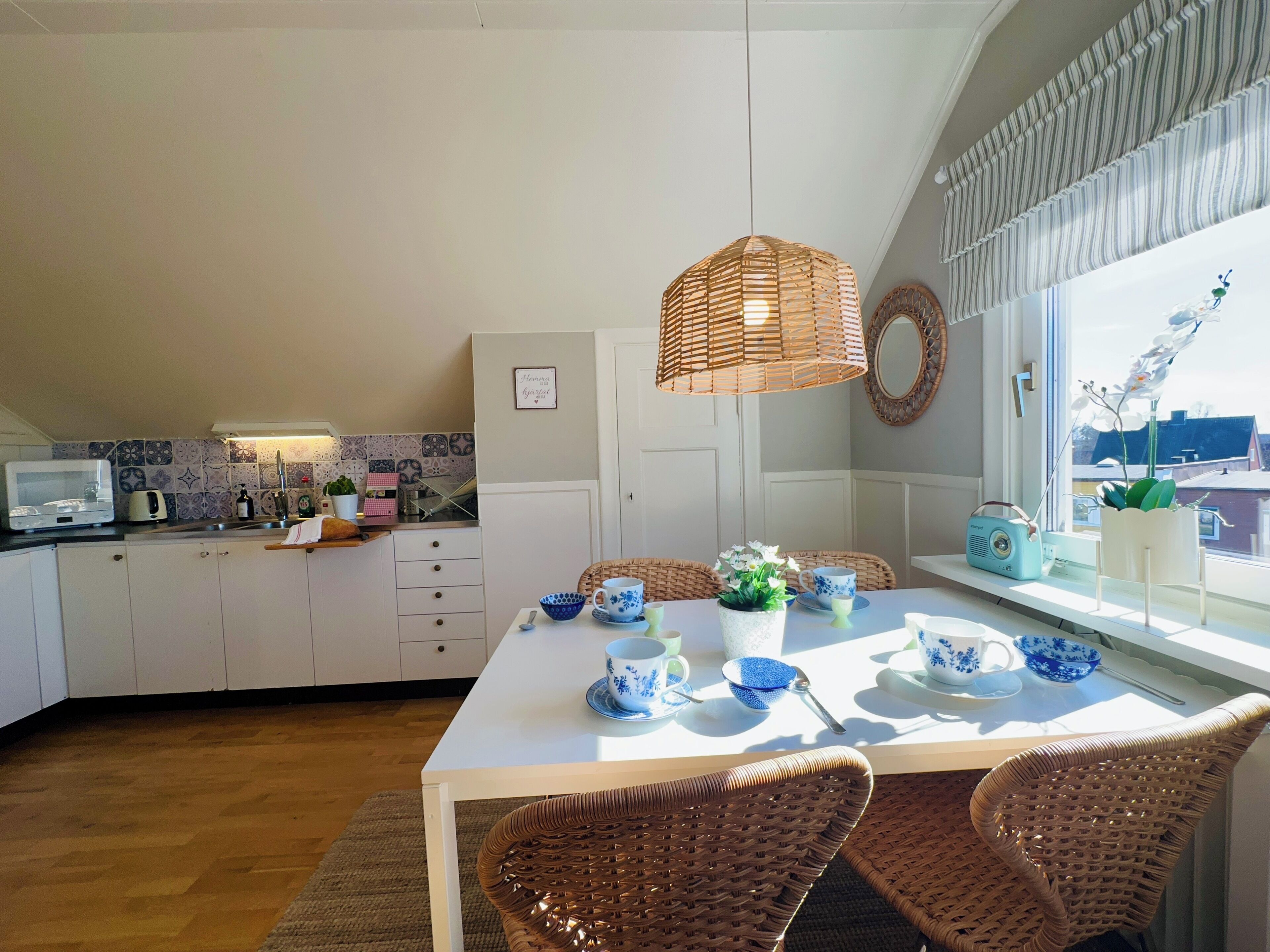 A kitchen with white cabinets, a dining table with blue and white patterned tableware, wicker chairs, a hanging wicker light fixture, and a window with a view of buildings.