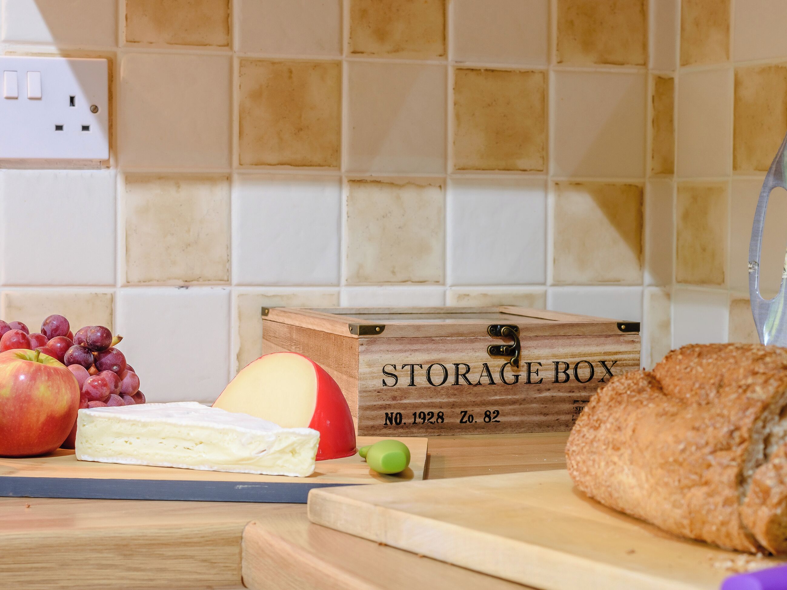 A kitchen counter with a wooden storage box labeled "STORAGE BOX," a cutting board, a loaf of bread, a halved apple, a wedge of cheese, and a bunch of grapes.