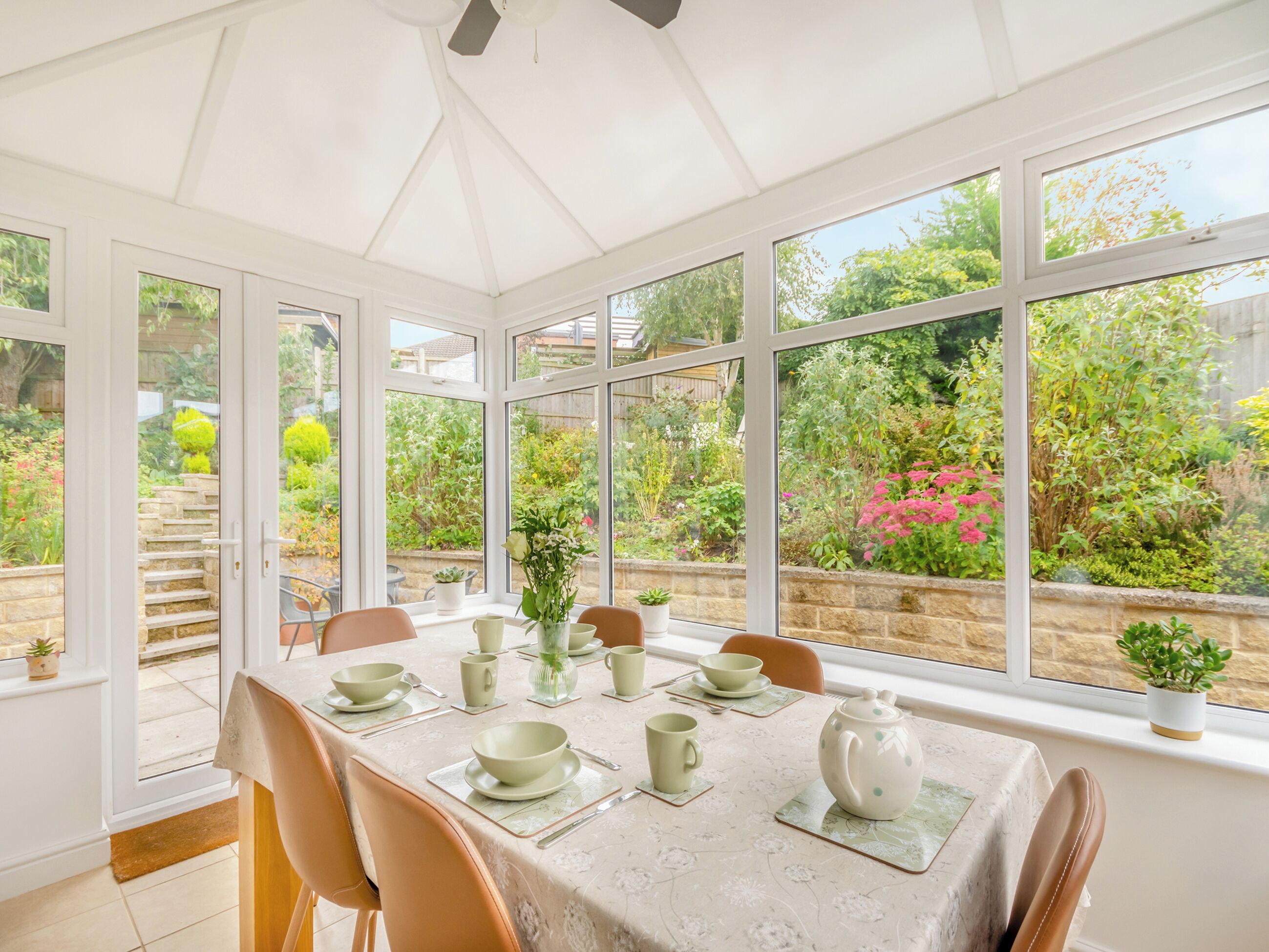 A conservatory with a table set for tea, surrounded by large windows offering a view of a garden.