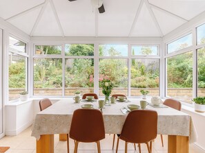 Un invernadero con una mesa dispuesta para comer, rodeado de amplios ventanales que dan a un jardín.