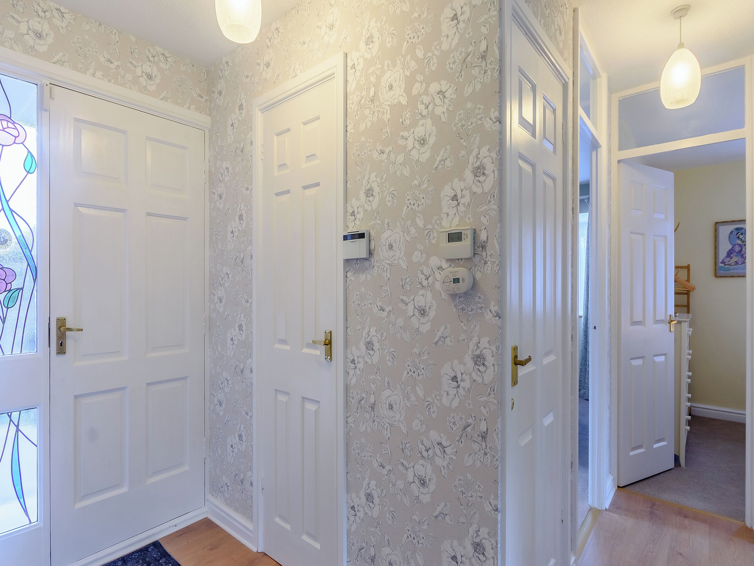 A hallway with white doors, floral wallpaper, a thermostat, and a light fixture.