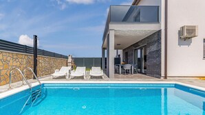 Et moderne poolområde med et klart, blåt pool, liggestole og en terrasse med et bord og stole.