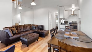 Uma sala de estar moderna com um sofá seccional, uma mesa de jantar com jogos americanos e uma cozinha com eletrodomésticos.