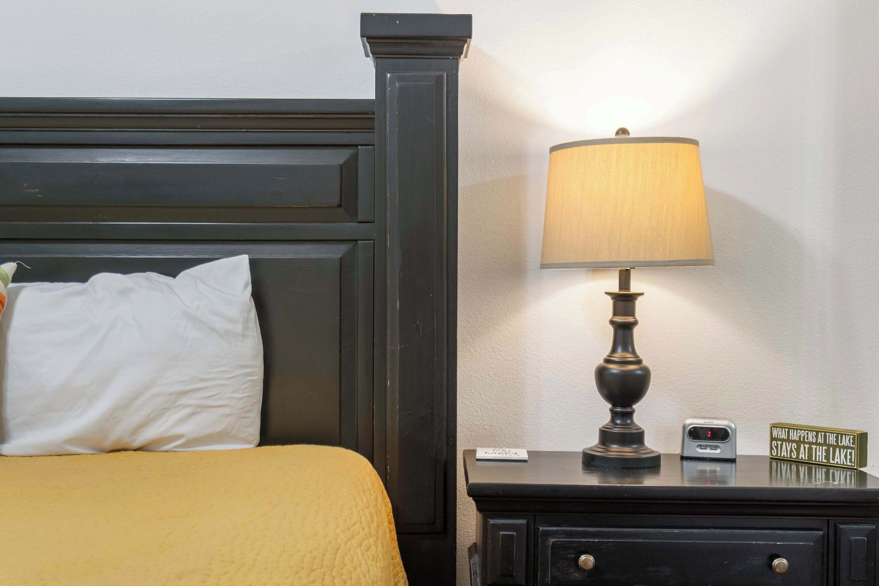 A bed with a white pillow and a yellow blanket, a dark wooden headboard, a bedside table with a lamp, a clock, and a sign that reads "WHAT HAPPENS AT THE LAKE, STAYS AT THE LAKE!".