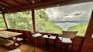 Terraza de madera con mesa y sillas, con vista a un lago y colinas boscosas.