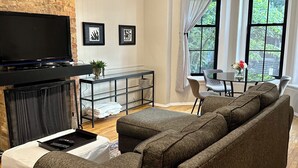 Uma sala de estar com sofá seccional, televisão de tela plana, mesa de centro de vidro e área de jantar com mesa e cadeiras.
