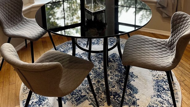 Quarto com uma mesa redonda preta, duas cadeiras estofadas cinzas, um vaso de vidro com flores e vista através de grandes janelas.