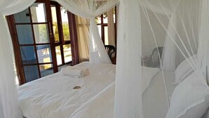 Un letto con biancheria bianca e un baldacchino, una finestra con vista sul verde, e una sedia sullo sfondo.