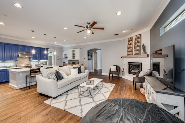 A spacious living area with a sofa, coffee table, and fireplace. A kitchen with blue cabinets and a dining area are visible in the background.