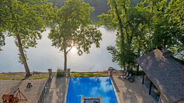 A swimming pool with a view of a lake, surrounded by trees and a small deck area with seating. - New Lakeside Haven - Picturesque (Branson)