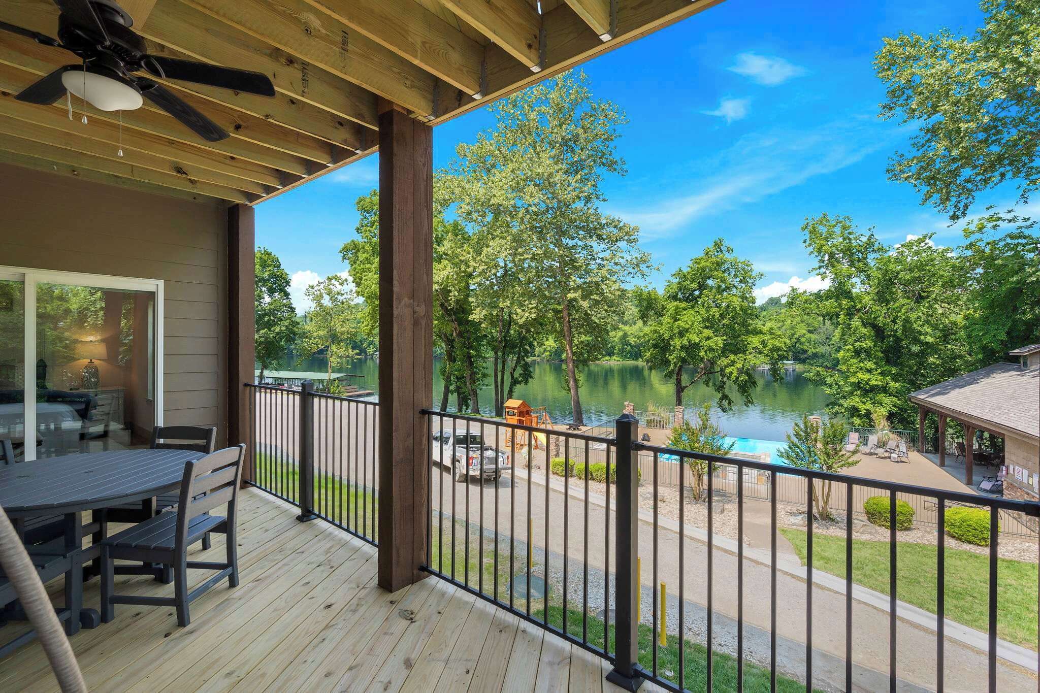 Terraza de madera con mesa y sillas, con vista a un lago que tiene un parque infantil y un muelle para barcos.